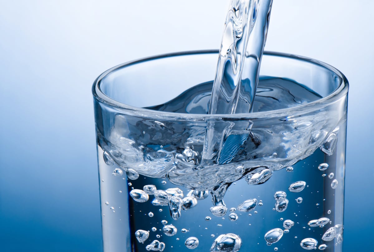 Agua cristalina cayendo dentro de un vaso transparente sobre fondo azul claro, con ondas y luz limpia.